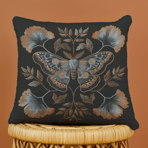 A square decorative pillow featuring an intricately detailed Luna moth in copper, terra cotta and slate blue colors on a black background. The moth is centered in a symmetrical arrangement with ginkgo-inspired leaves and botanical elements surrounding it.